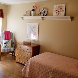A cozy bedroom with simple decor, featuring a bed, a wooden dresser, a chair by the window, and artwork on the walls.