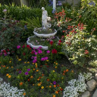 A beautiful garden scene featuring a small fountain with a statue surrounded by vibrant flowers and lush greenery.