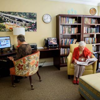 An elderly man using a computer and an elderly woman reading in a cozy library setting.