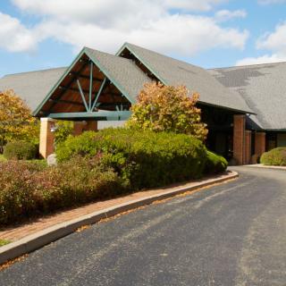 A modern building with a sloped roof surrounded by lush greenery and a curved driveway under a partly cloudy sky.