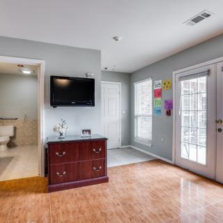 A cozy living area featuring a wall-mounted TV, wooden dresser, and bright artwork on the walls. The room connects to a bathroom and has a glass patio door letting in natural light.