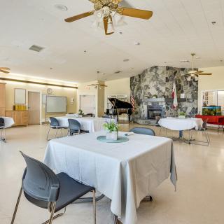 A spacious and well-lit dining area featuring tables with white tablecloths, modern chairs, a grand piano, and a stone wall.