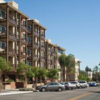 A modern apartment complex on a sunny street lined with parked cars and palm trees.