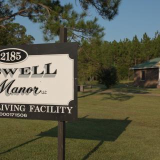 Sign for Maxwell Manor, a living facility, located in a grassy area with trees and a building in the background.