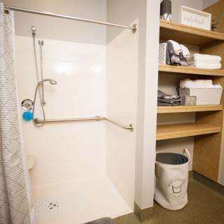 A bathroom with a walk-in shower equipped with grab bars, adjacent shelving holding towels and toiletries, and a small trash bin.