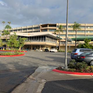A view of a modern hospital building with a parking lot in the foreground, surrounded by greenery under a cloudy sky.