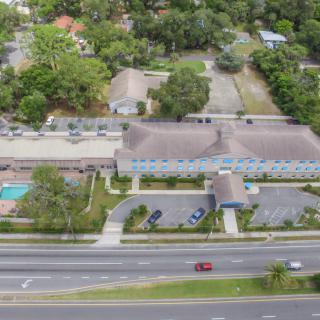 An aerial view of a building complex with a road in front, parking lots, and a swimming pool surrounded by trees.