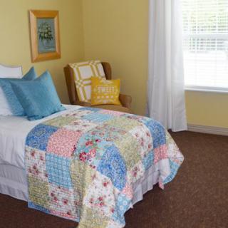 A cozy bedroom with a single bed featuring a colorful patchwork quilt and decorative pillows, next to an armchair by the window.