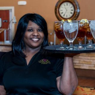 A smiling waitress holding a tray of iced tea glasses in a restaurant setting.