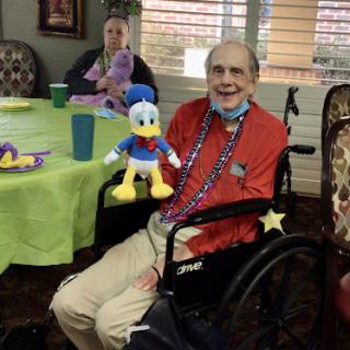 An elderly man in a wheelchair happily holds a plush Donald Duck toy during a social gathering at a residential facility.