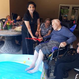 An elderly person relaxes with their feet in a small pool, assisted by a caregiver, as others enjoy the moment at a care facility.