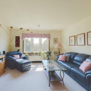 A cozy living room with a leather sofa, armchair, and coffee table, featuring artwork on the walls and natural light from a large window.