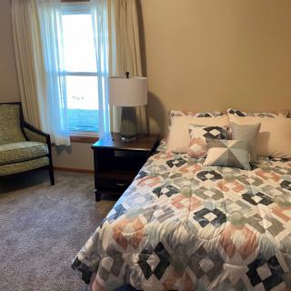 A cozy bedroom featuring a neatly made bed with patterned bedding, a nightstand with a lamp, and a window with curtains.