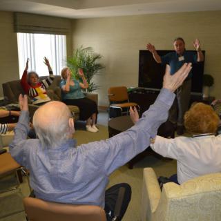 A group of elderly individuals participating in an indoor exercise session, raising their arms as part of the activity.