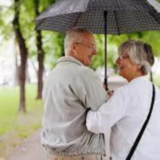 An elderly couple enjoys a walk together under an umbrella along a tree-lined path.