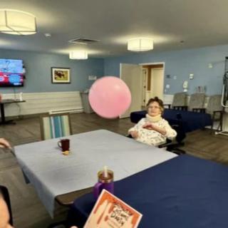 An elderly woman sitting in a recreation room, playing with a pink balloon over a table.