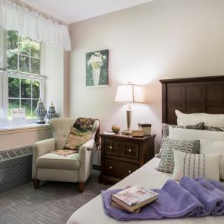 A cozy bedroom with a neatly made bed, comfortable armchair, and a side table with a lamp.