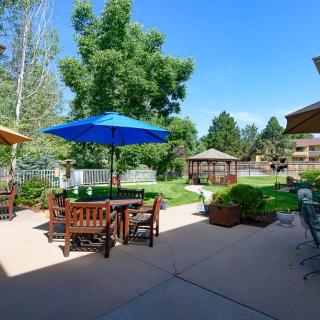 A sunny outdoor patio with wooden furniture and a blue umbrella, surrounded by trees and a gazebo in the background.