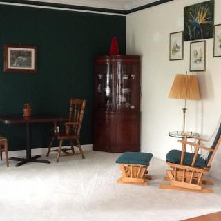 A cozy living room with dark green and white walls, featuring wooden furniture including chairs and a table, a cabinet, floor lamp, and various wall decorations.