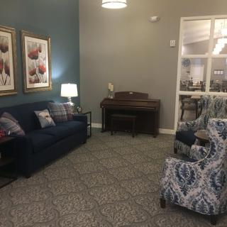 A cozy living room with a sofa, armchair, and piano, adorned with framed art.