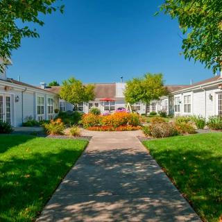 A scenic view of a garden pathway leading to a building, surrounded by lush greenery and vibrant flowers under a clear blue sky.