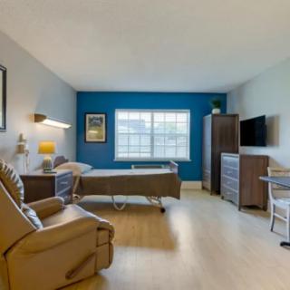 A well-furnished bedroom featuring a bed, armchair, television, and large window with natural light.