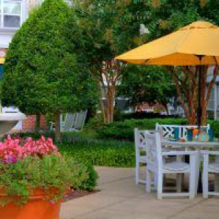 A serene patio setting with a table, chairs, and a yellow umbrella surrounded by lush greenery and colorful flowers.