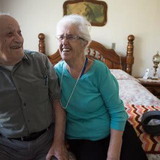 An elderly couple smiling and sitting together in a cozy bedroom.