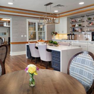 A modern kitchen with an island counter and a cozy dining area featuring elegant chairs and a wooden floor, accented by a hanging chandelier and fresh flowers.