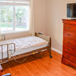A bright bedroom with a single hospital bed next to a window, featuring a wooden dresser and a TV.