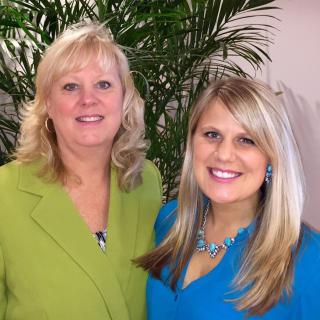Two women smiling indoors with a plant in the background.