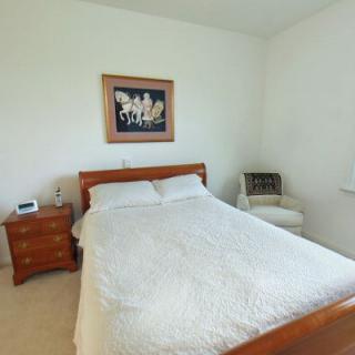 A minimalist bedroom with a neatly made bed, wooden nightstand, artwork on the wall, and a chair in the corner.