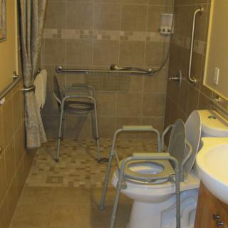An accessible bathroom equipped with grab bars, a commode chair, and a shower area.