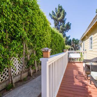 A sunny deck alongside a house with lush greenery and a white fence.