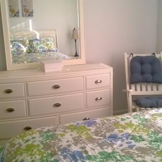 A cozy bedroom featuring a white dresser with a mirror, a cushioned chair, and a bed with a floral patterned bedspread.
