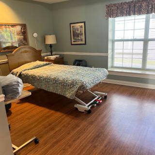 A spacious hospital room featuring a patient bed, window with natural light, and warm wooden flooring.