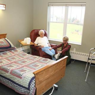 An elderly person sitting in an armchair next to a hospital bed in a nursing home room.