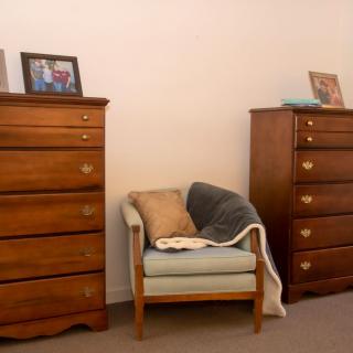 A cozy room setup with two wooden dressers and a cushioned chair in between, adorned with cushions and a blanket.