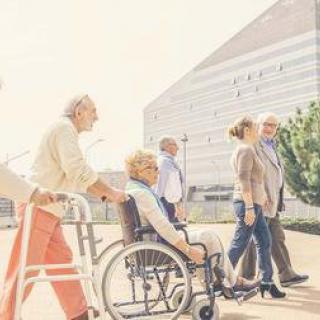 A group of elderly people enjoying a walk outdoors, with one person using a wheelchair and another using a walker.