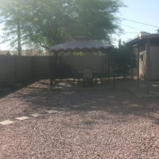 A sunny backyard with gravel ground, featuring a gazebo and outdoor seating area surrounded by trees.