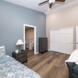 A cozy bedroom features a blue accent wall, wooden flooring, and simple furnishings including a bed, nightstand, dresser, and ceiling fan.