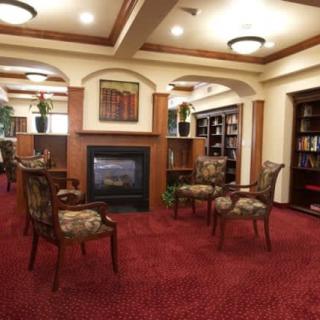 A cozy library interior with a fireplace, surrounded by bookshelves and chairs on a carpeted floor.