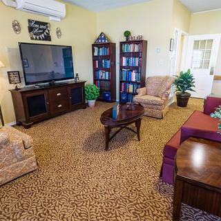 A cozy living room featuring a large TV, armchairs, bookshelves, and a coffee table on a patterned carpet.
