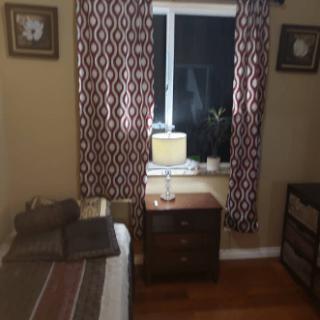 A cozy bedroom featuring a bed, nightstand with a lamp, and patterned curtains framing a window.