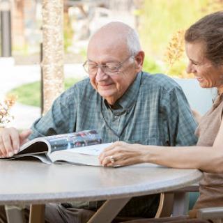 An elderly man and a woman enjoy reading together outdoors, sharing smiles over a magazine.