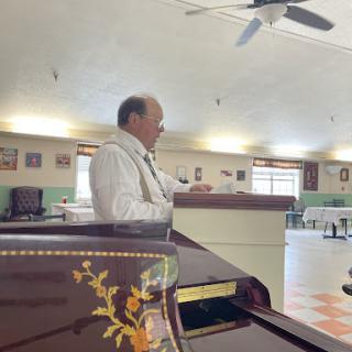 A man speaks in a spacious interior, with a piano partially visible in the foreground.