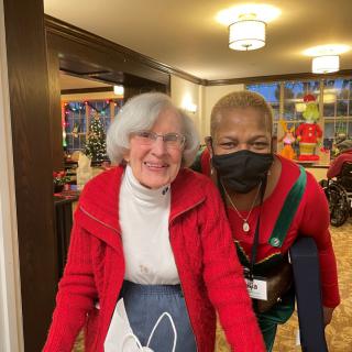 An elderly woman and her caregiver share a moment together indoors, both wearing red clothing.