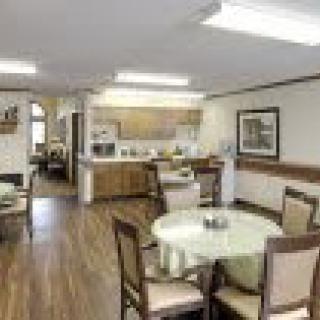 A cozy dining area with several tables and chairs, connected to a kitchen with wooden flooring and warm lighting.