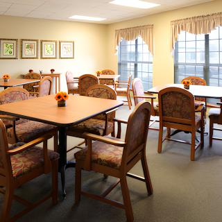 A cozy dining area with wooden tables and chairs, decorated with small centerpieces, featuring large windows with curtains.