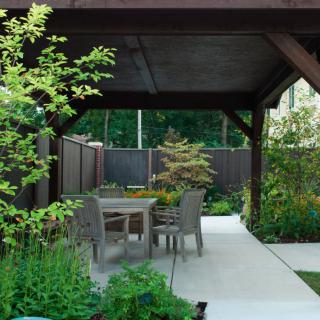 A tranquil garden patio with a wooden pergola and outdoor seating surrounded by lush greenery.